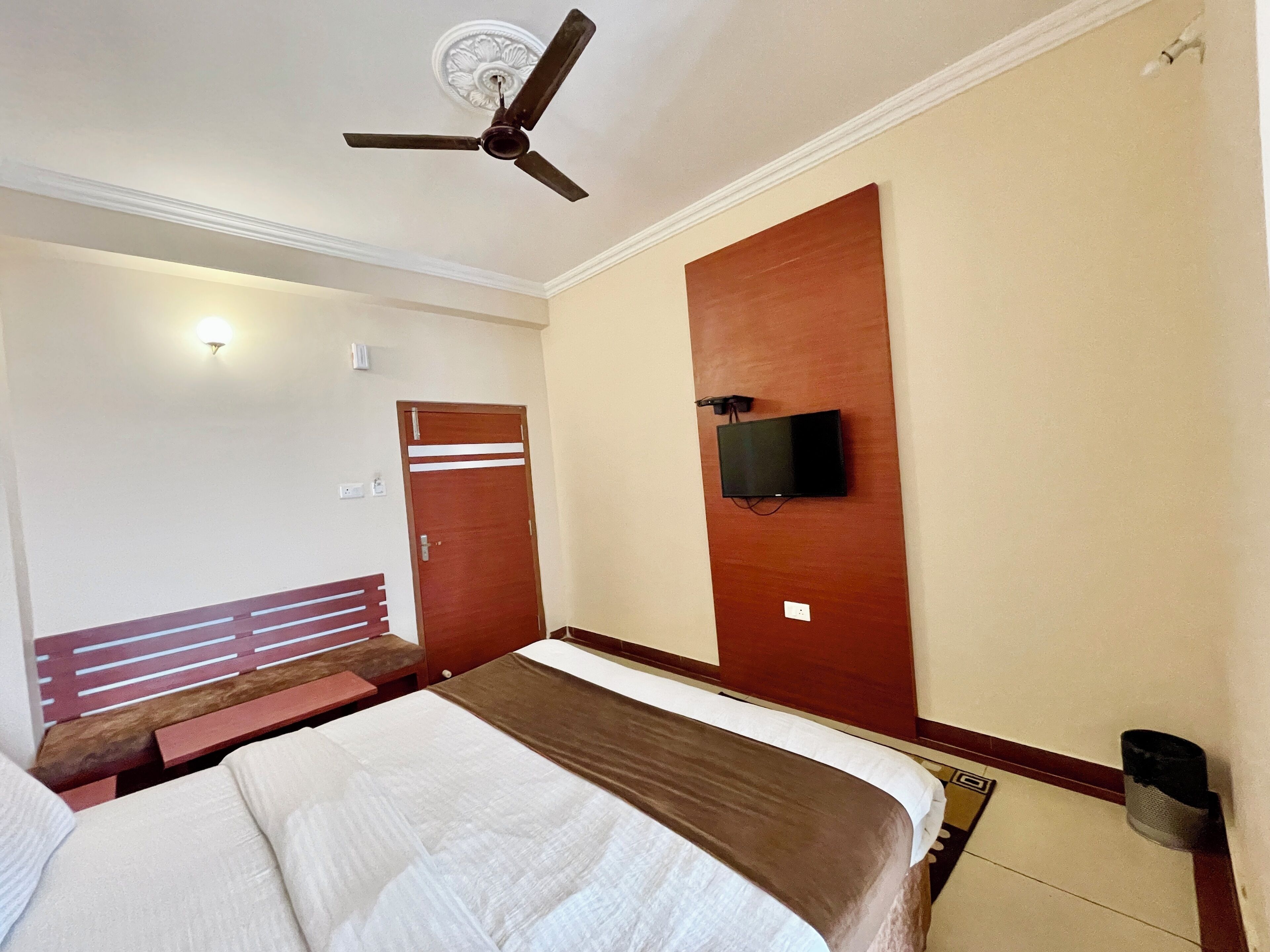 A hotel room with a bed, a ceiling fan, a television mounted on the wall, and a wooden bench.