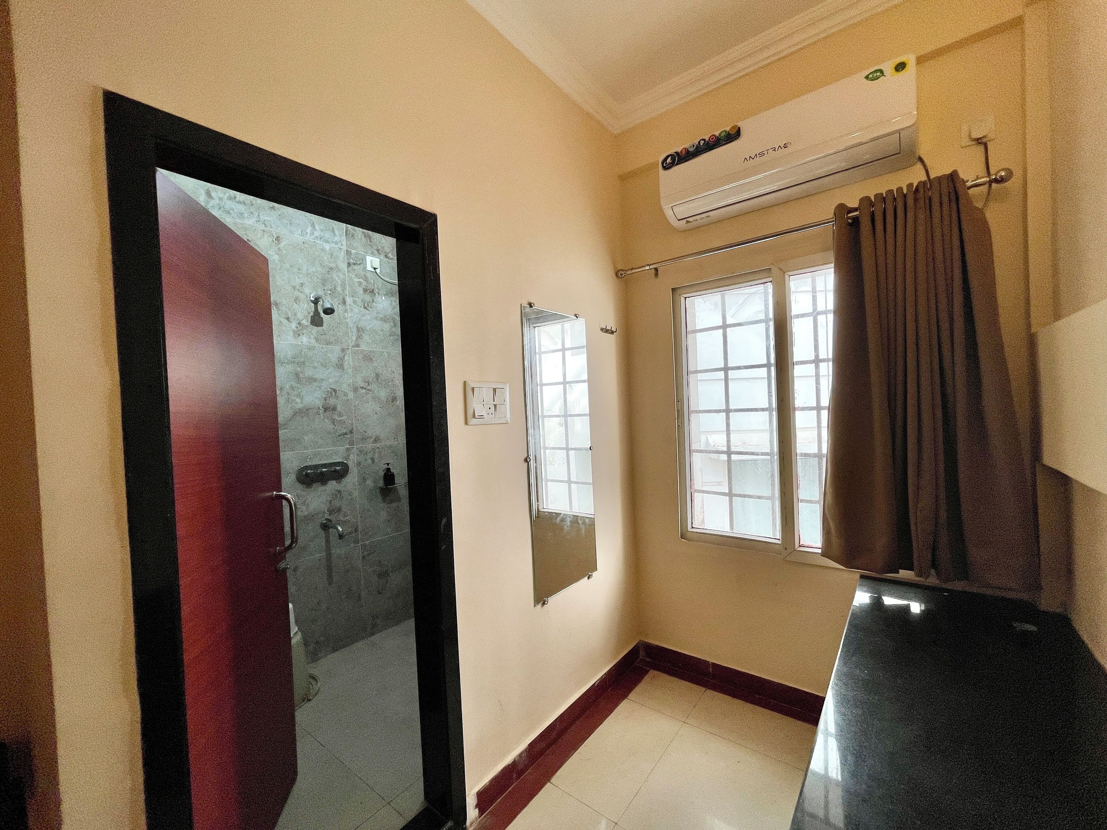 A bathroom with a shower, a mirror, a window with curtains, and an air conditioning unit.