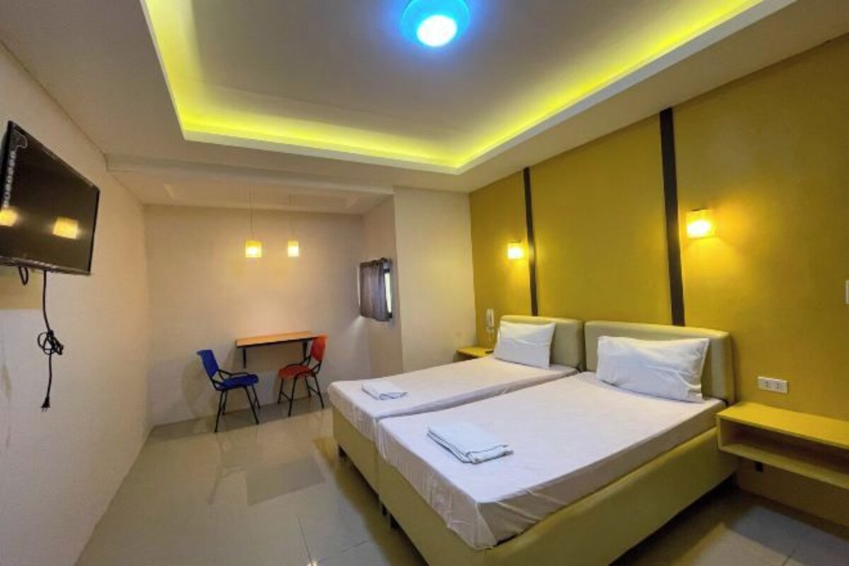 A hotel room with two beds, a desk, a chair, a TV, and a ceiling light.