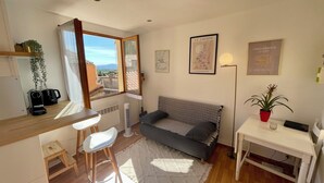 Un espace de vie compact comprenant un canapé, une petite table, une machine à café, un radiateur et une fenêtre donnant sur un paysage agréable.