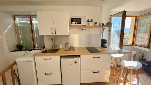 Une cuisine compacte avec des meubles blancs, un four à micro-ondes encastré et un petit réfrigérateur. On y trouve des tabourets et une fenêtre donnant sur l’extérieur.