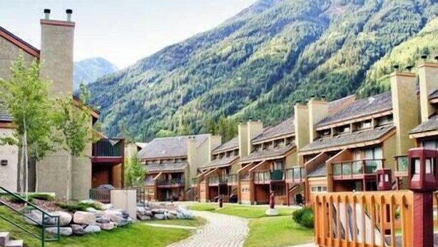 A row of chalet-style buildings with a stone facade, a central pathway, and a mountainous backdrop. - Ski In/Out - Hot tub - Fireplace - BBQ (Panorama)