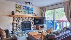 A living room with a stone fireplace, a flat-screen TV, a leather couch, and a large window with a view of a playground. - Ski In/Out - Hot tub - Fireplace - BBQ (Panorama)