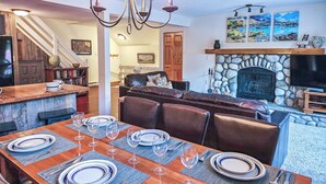 A dining area with a wooden table set for six, a stone fireplace, and a chandelier. - Ski In/Out - Hot tub - Fireplace - BBQ (Panorama)