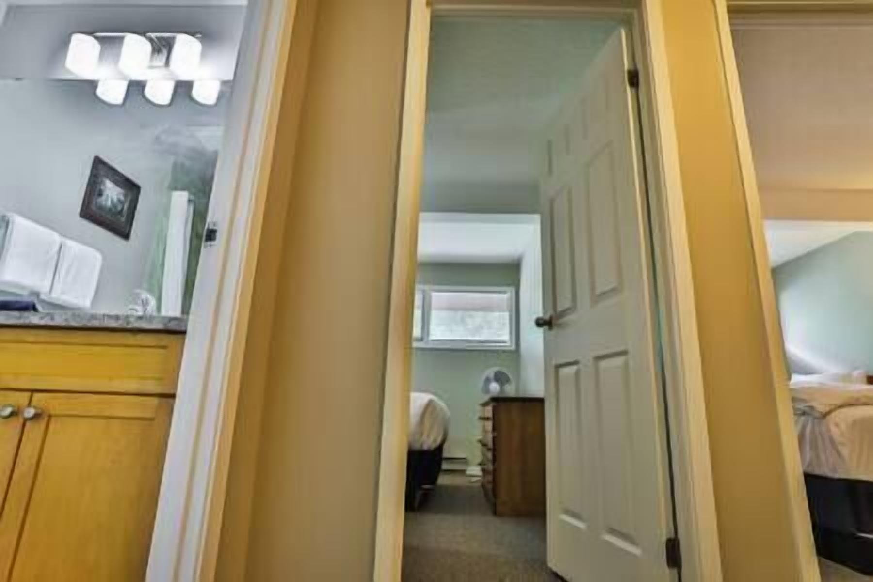 A narrow hallway leading to a bedroom with a bed and a bedside table. There is a door on the right and a window with blinds in the background.