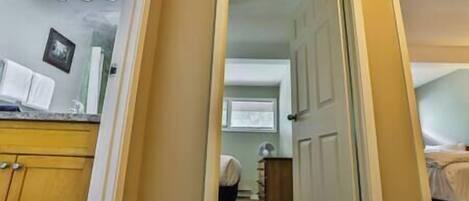 A narrow hallway leading to a bedroom with a bed and a bedside table. There is a door on the right and a window with blinds in the background.