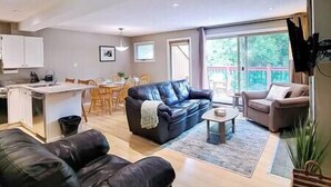 A modern living room with a black leather sofa, a beige armchair, and a coffee table. There is a dining area with wooden chairs and a white table. The room has large windows and a view of greenery outside. - New Beds / AC / Pool & Hot Tub / Fireplace (Panorama)