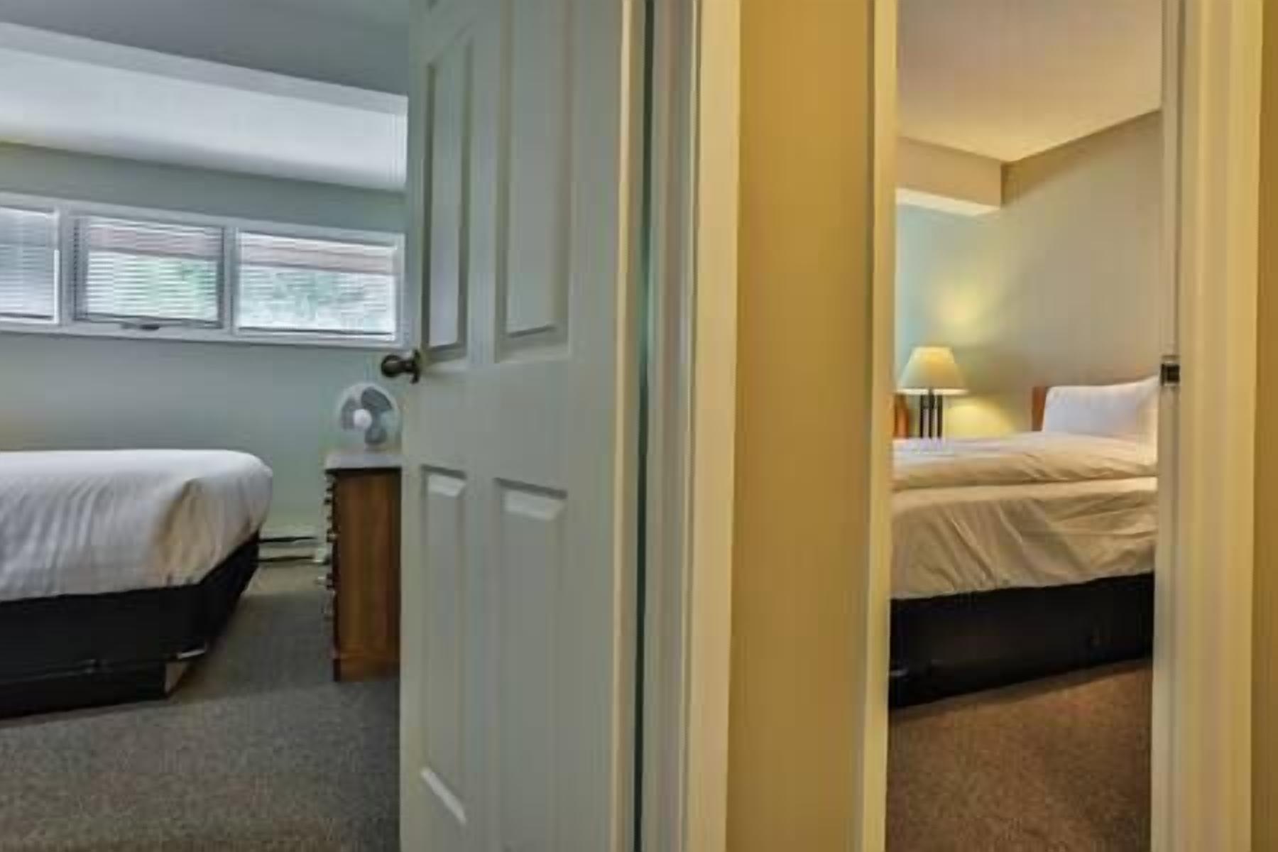 A hotel room with two beds, a bedside table, a lamp, and a window with blinds.