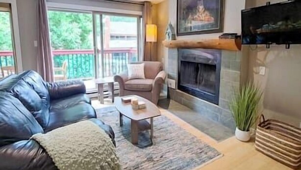 A living room with a leather sofa, a coffee table, a fireplace, and a balcony with a view of greenery. - New Beds / AC / Pool & Hot Tub / Fireplace (Panorama)