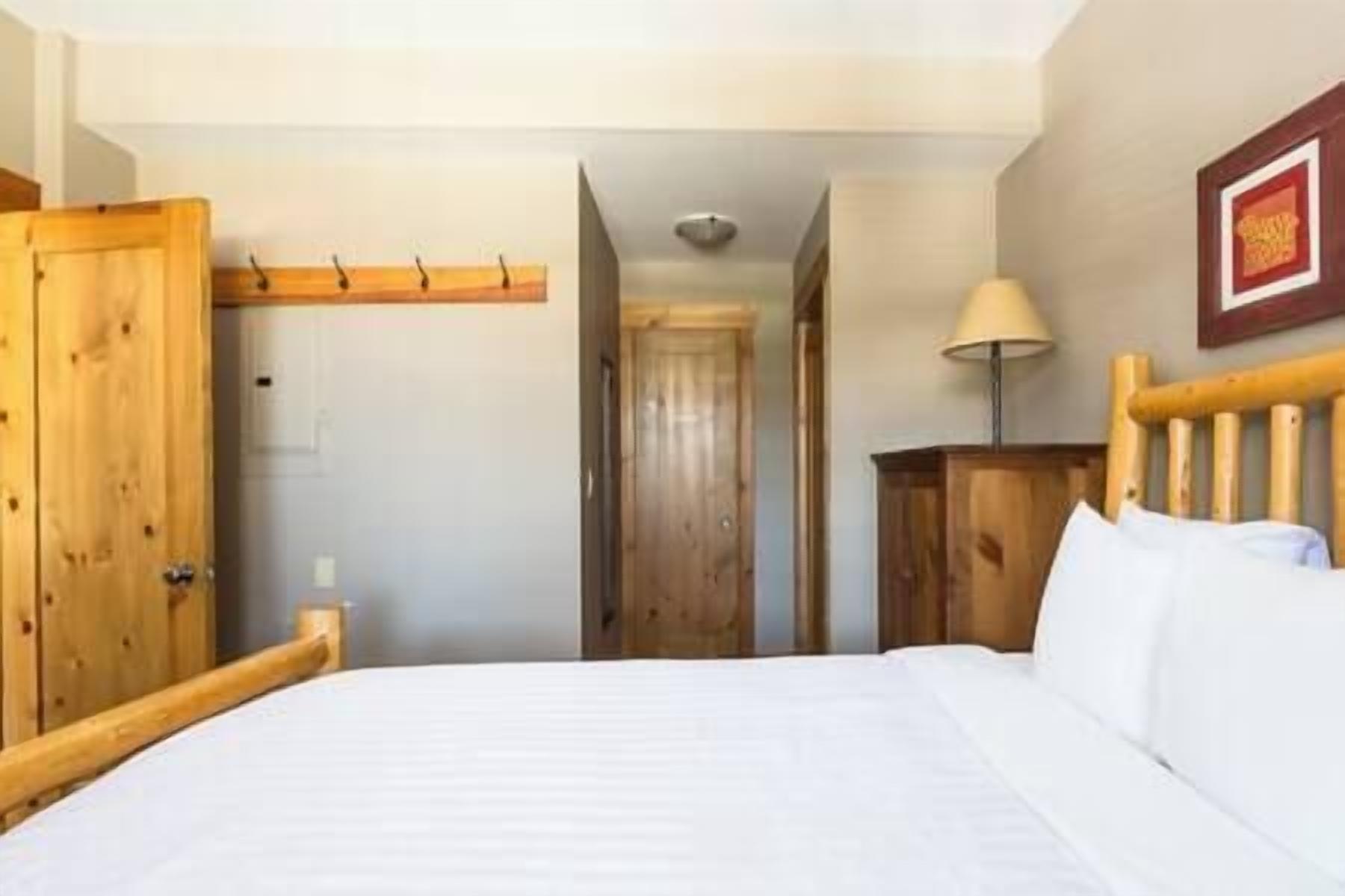 Habitación de hotel con cama, cabecera de madera, armario de madera, lámpara y un cuadro en la pared.