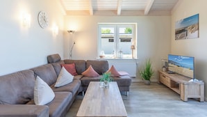 Ein Wohnzimmer mit braunem Sofa, einem Holztisch, einem Fernseher auf einem Holzboden, einer Pflanze und einem Fenster mit Blick ins Freie.