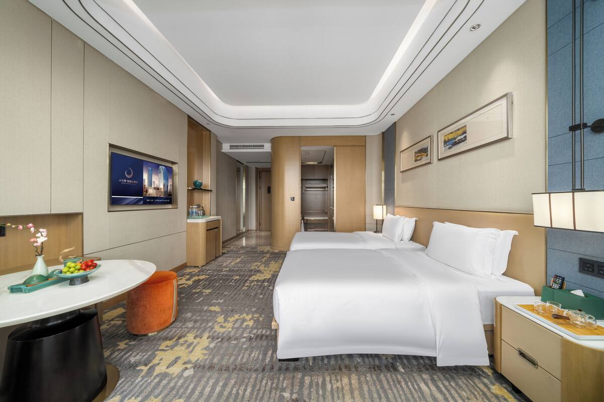 Una habitación de hotel con una cama grande, un escritorio con un florero, un cesto de basura y piso alfombrado.