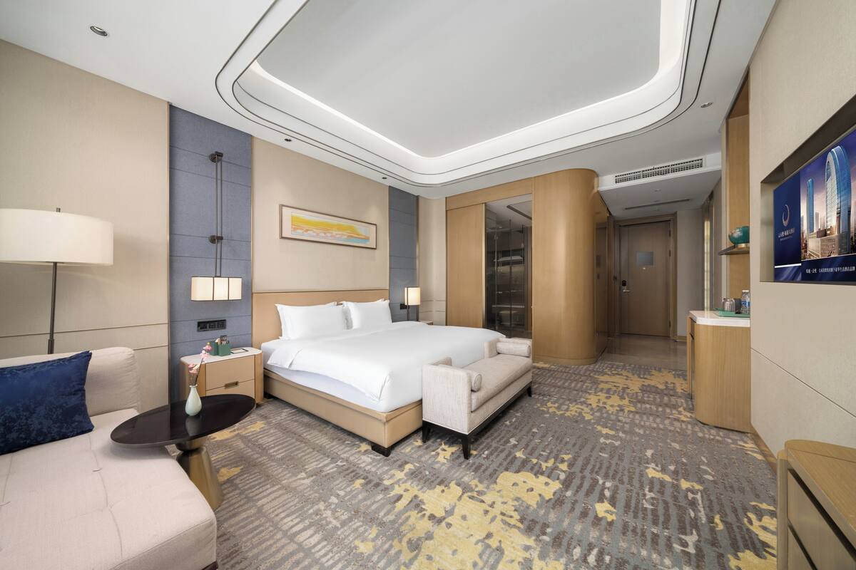 Habitación de hotel con una cama grande, un sofá, un escritorio y un baño visible a través de una puerta de cristal.