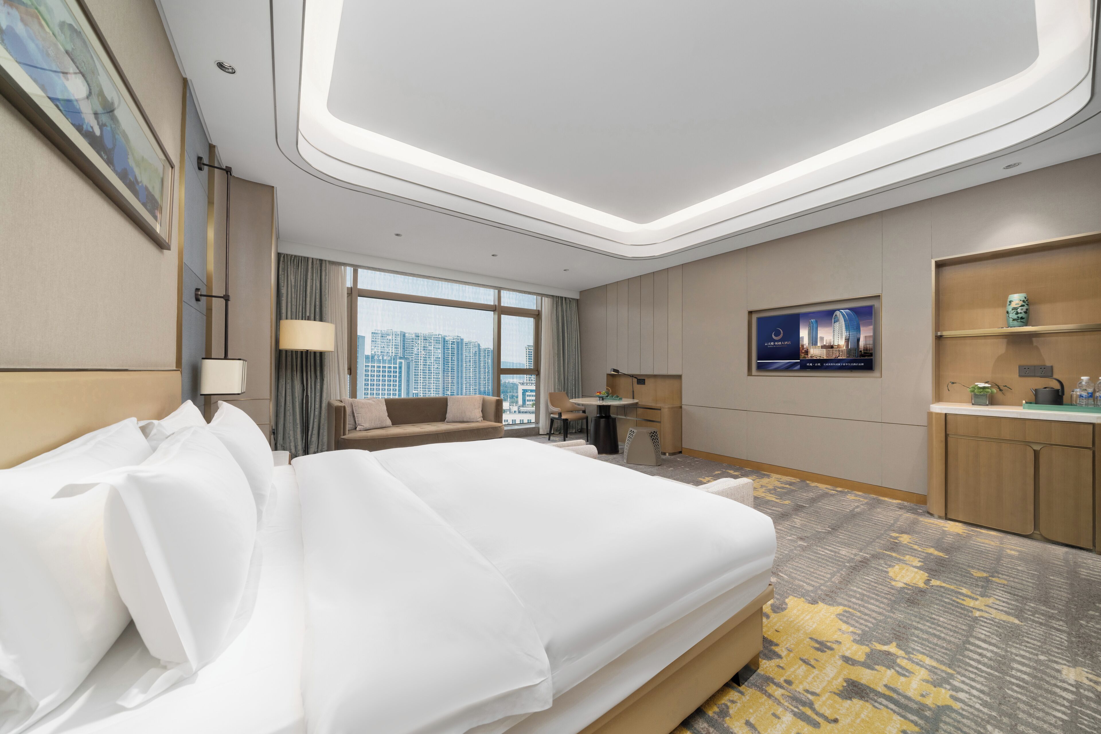 Habitación de hotel con una cama grande, un escritorio, una silla, un sofá, un ventanal con vista a la ciudad y una pequeña cocina americana.