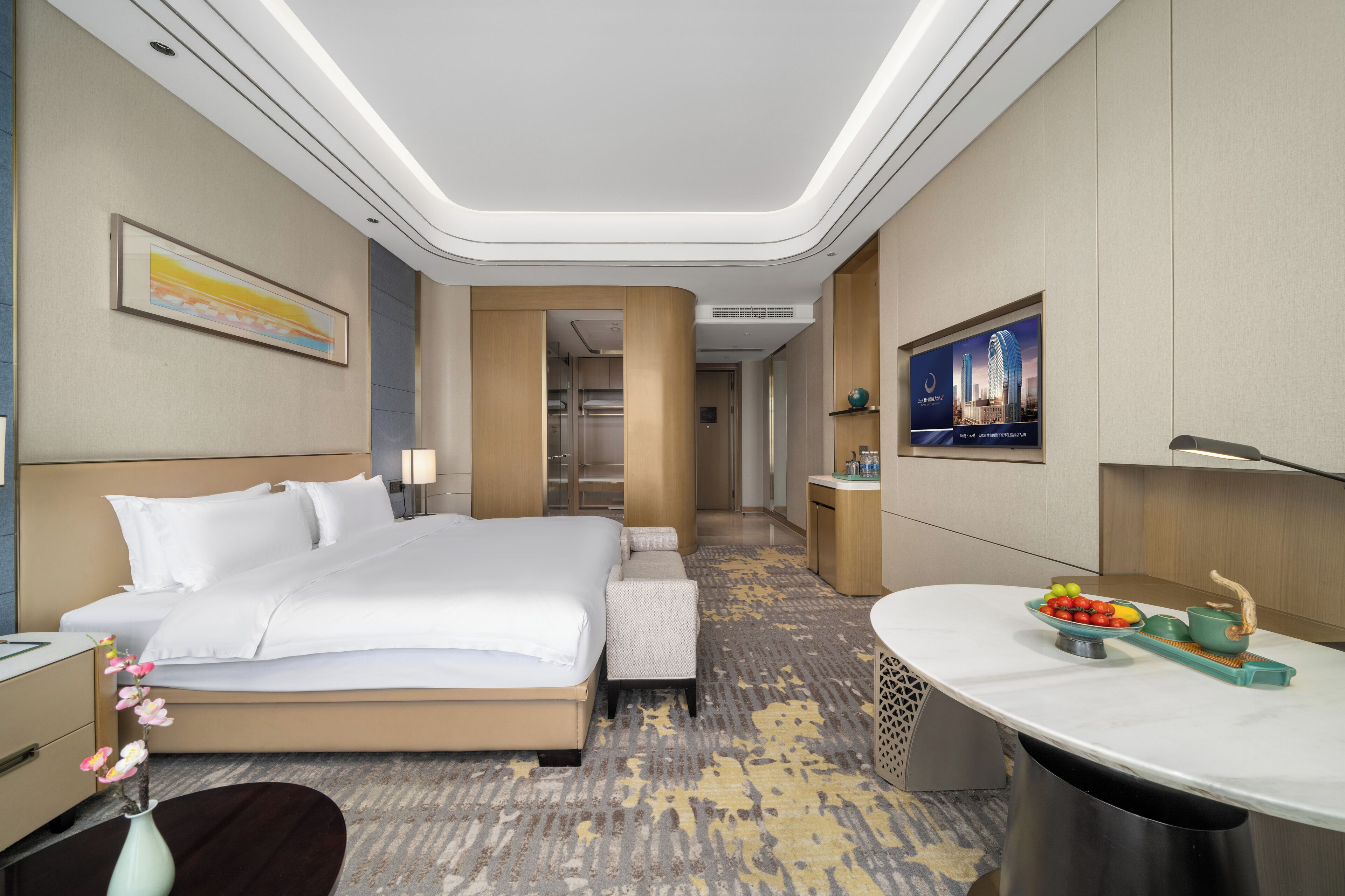 Une chambre d’hôtel avec un grand lit, un bureau sur lequel se trouve un panier de fruits et un téléviseur fixé au mur.