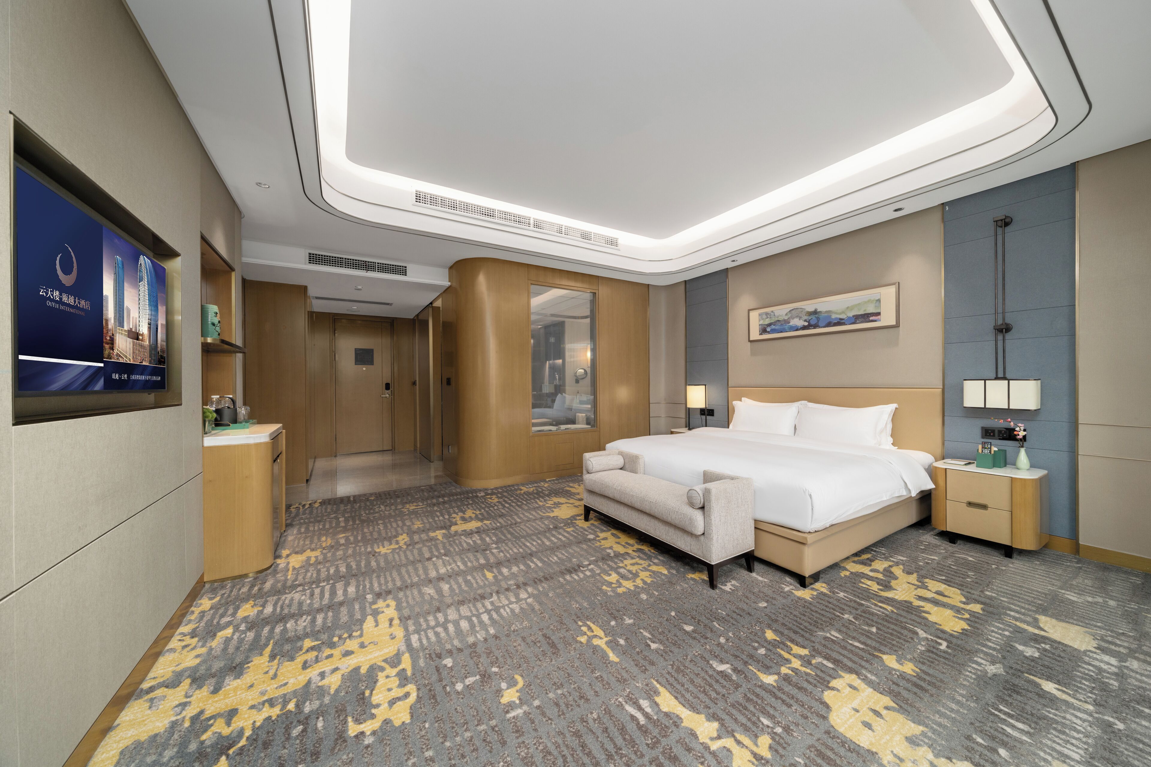 A hotel room with a large bed, a nightstand, a desk, a chair, a sofa, a carpet, a painting, a television, and a poster on the wall.