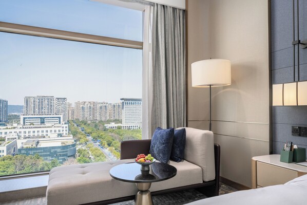 A hotel room with a sofa, a small round table with a fruit bowl, a lamp, and a view of a cityscape.