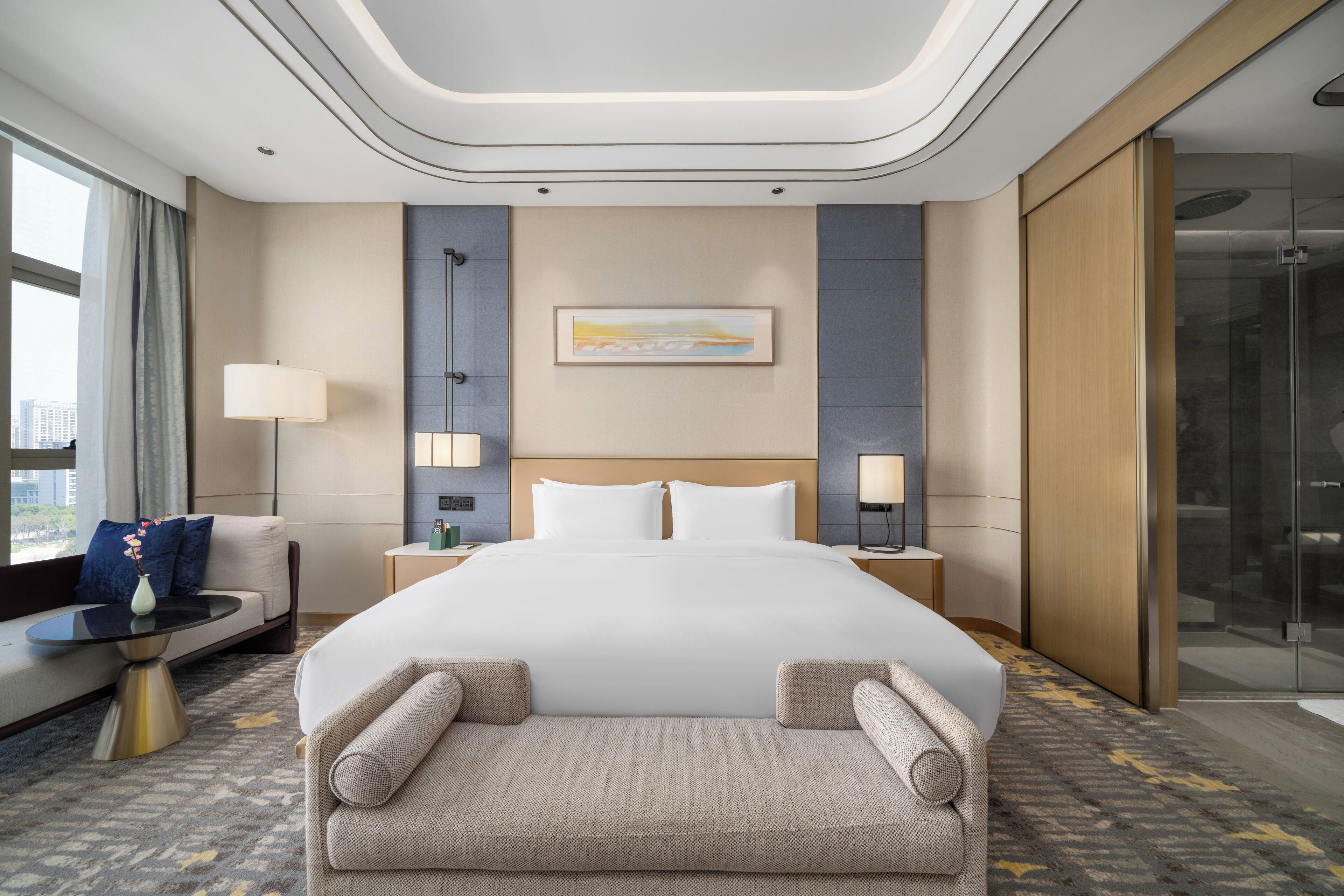 A modern hotel room with a large bed, a sofa, a small table, and a glass shower area.