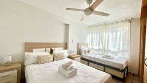 A hotel room with a large bed, a smaller bed, a ceiling fan, a nightstand, and a window with sheer curtains. - Sunrise at Home by Gloove (El Campello)