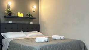 Ein ordentlich bezogenes Bett mit einer grauen Decke, zwei weißen Kissen und zwei aufgerollten Handtüchern. Darauf befindet sich ein Regal mit einer Topfpflanze und zwei Kerzen.