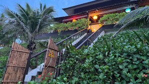 A staircase leading to a building with a traditional roof, surrounded by lush greenery and a bamboo fence.