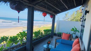 A covered outdoor seating area with a view of the beach, featuring a blue sofa, red cushions, and potted plants.