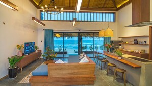 A modern living room with a large window overlooking the ocean, a wooden bench, a kitchen area with bar stools, and a TV on a console table.