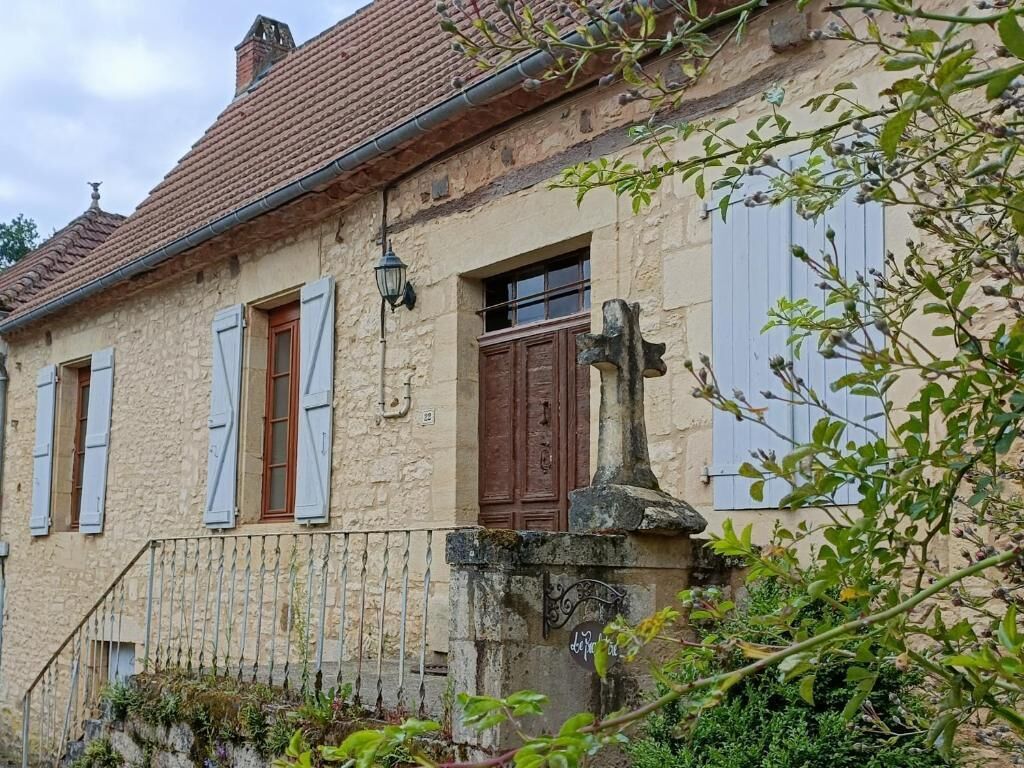 A rustic stone house with a tiled roof, a wooden door, and a small garden area with a metal fence.