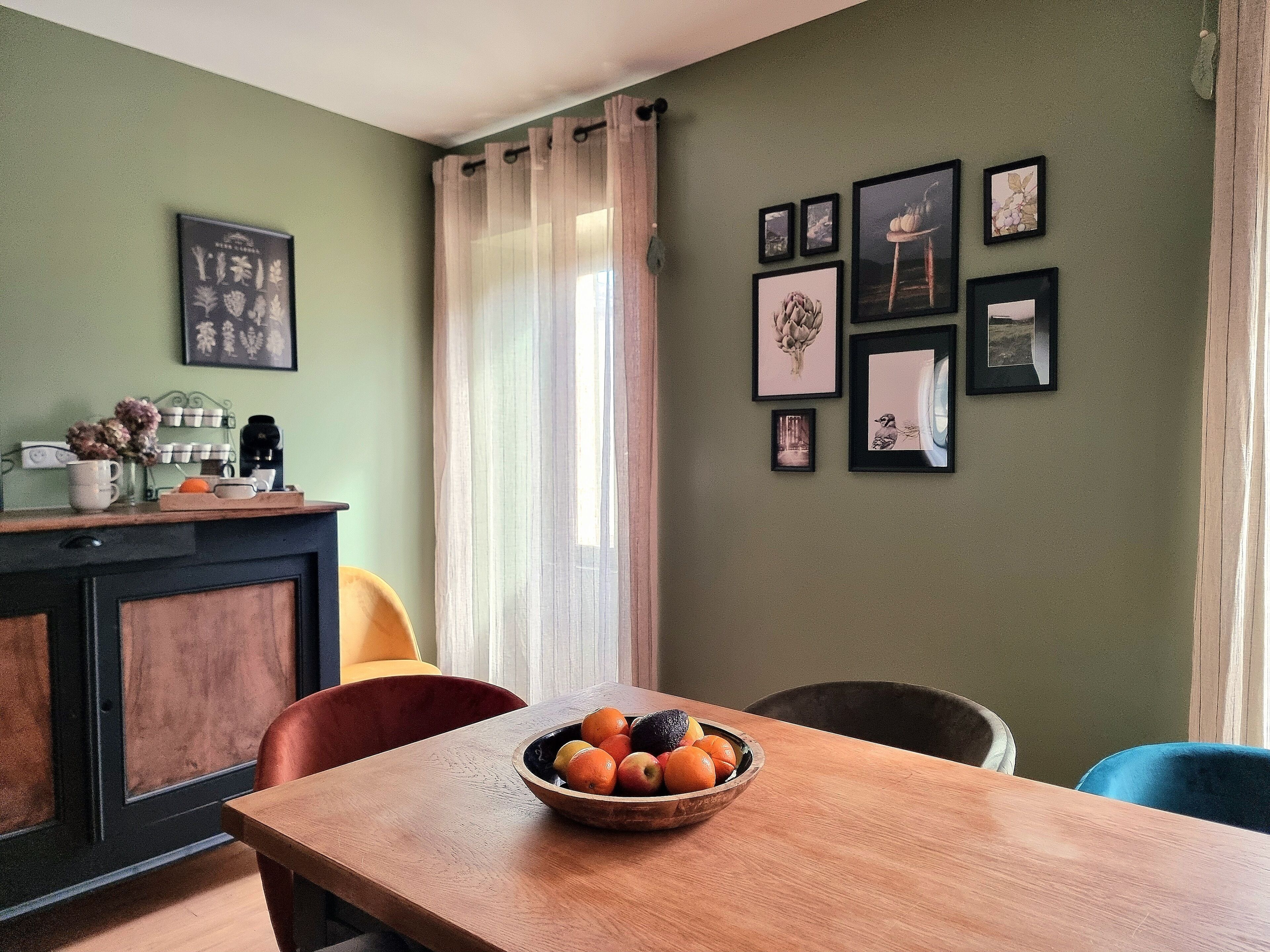 A dining area with a wooden table, chairs, and a bowl of fruit. There are framed pictures on the wall and a window with curtains.