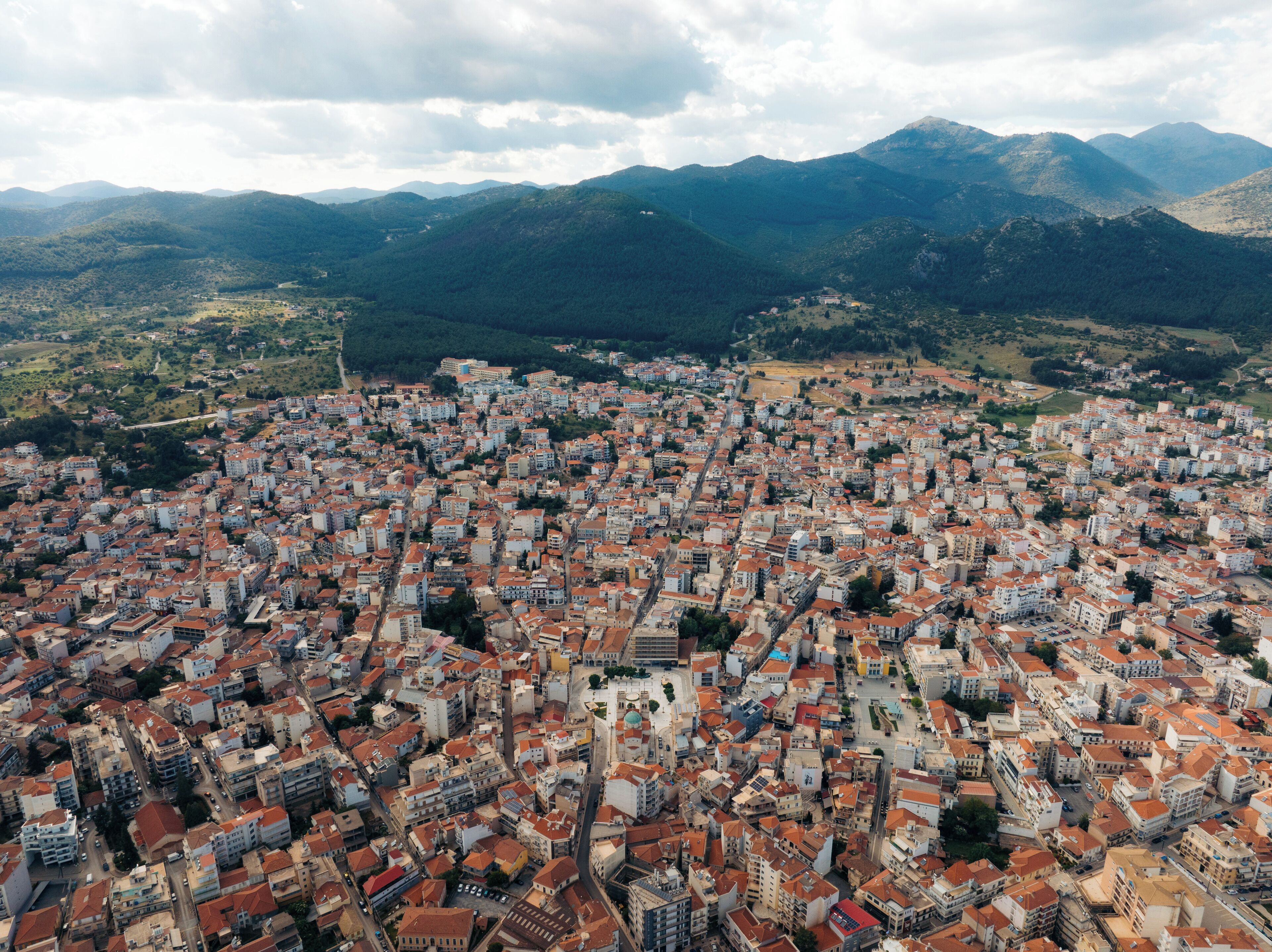 An aerial view of a densely packed urban area with red-roofed buildings, surrounded by green hills and mountains.