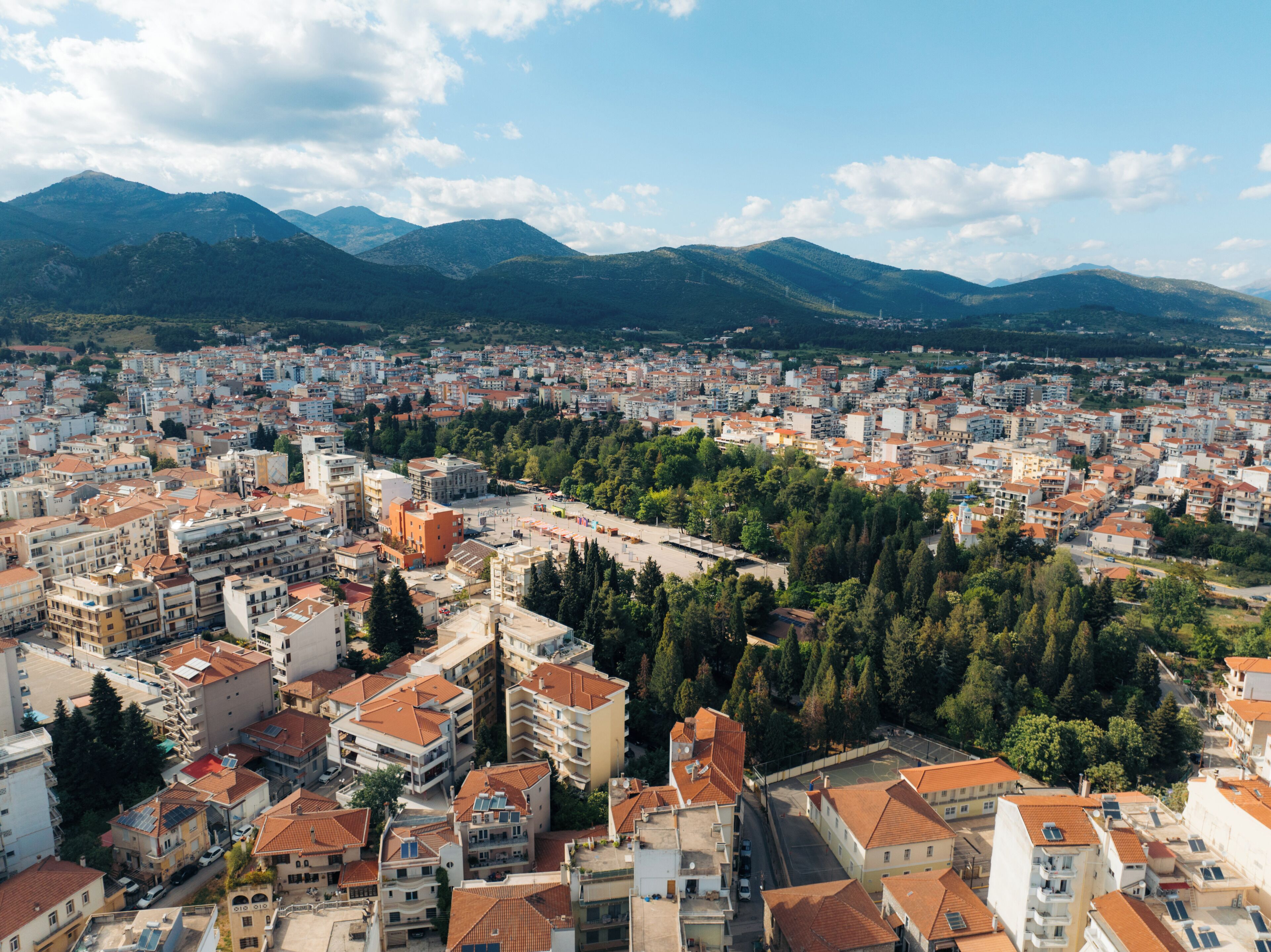 A cityscape with a dense cluster of buildings, some with red-tiled roofs, surrounded by greenery and mountains in the background.