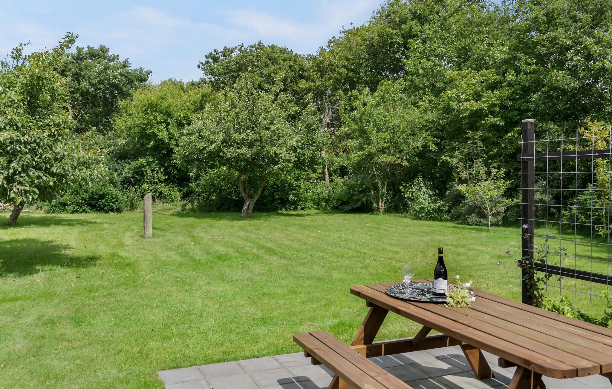 Ein Picknicktisch mit einer Weinflasche und Gläsern, auf einer Terrasse mit Blick auf einen üppigen Garten und Bäume.