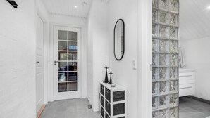 Un couloir avec une porte blanche, un miroir, une petite armoire et une cloison en verre.