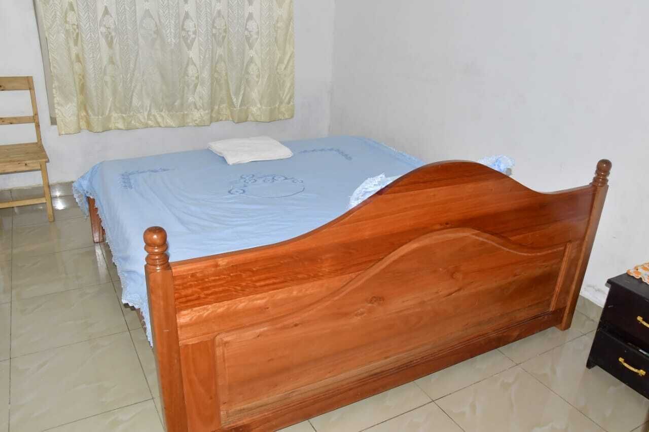 Una cama de madera con cubrecama azul, una mesita de noche de madera con cajones y una cortina en la ventana.