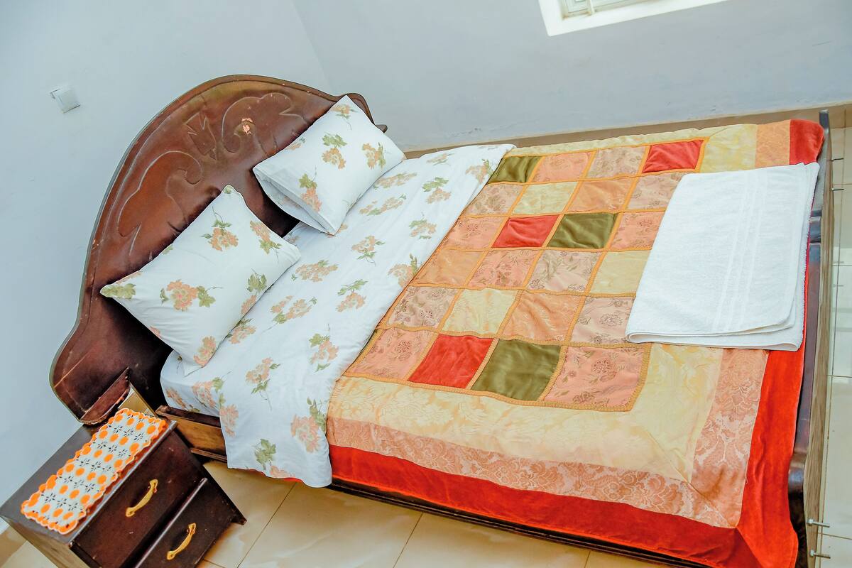 Una cama bien hecha con almohadillas florales y colcha, una mesita de noche de madera con una bandeja estampada, y una ventana que deja entrar luz natural.