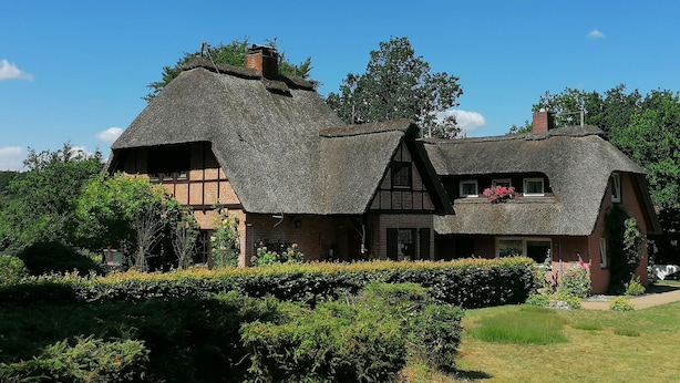 Une maison traditionnelle à toit de chaume, dont la partie inférieure est en briques, entourée de haies et d’arbres bien entretenus.