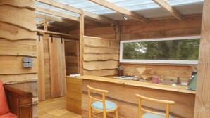 Interior de una cabaña de madera con una zona de cocina que incluye una encimera de madera, dos taburetes de bar, una ventana y una puerta que da a otra habitación.