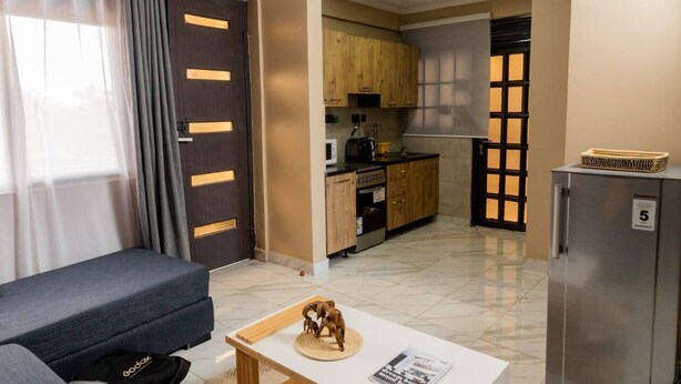 A compact living space with a sofa, a small table with a decorative item, a kitchenette with wooden cabinets, and a refrigerator. - Fed Stay Naalya (Kampala)
