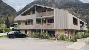 Un edificio moderno a due piani con balconi, un'area di parcheggio e montagne sullo sfondo.