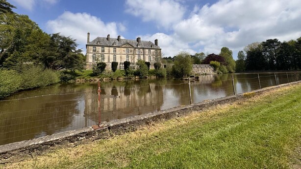 A large stone building with a river in front, surrounded by trees and a grassy area.