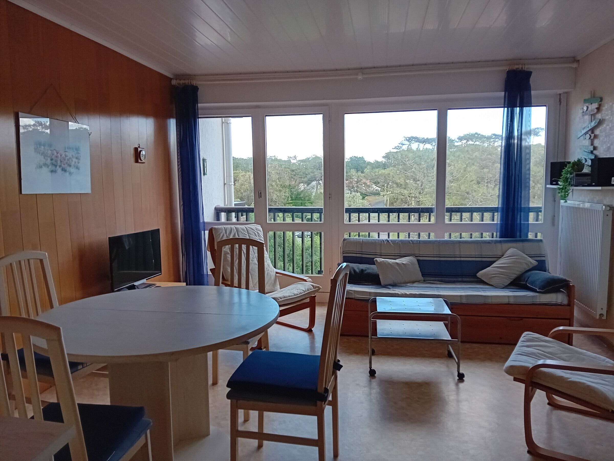 Ein Zimmer mit einem runden Tisch, Stühlen, einer Couch und einem Fernseher. Große Fenster bieten Blick auf Bäume und Hügel.
