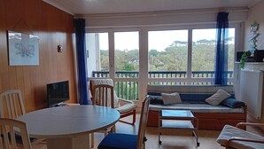 A room with a round table, chairs, a sofa, and a TV. Large windows offer a view of trees and hills. - Seaside Retreat: Cozy 1BR Apt Steps from Beach & Shops (Seignosse)