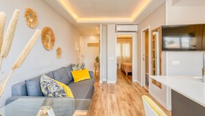 A modern living room with a grey sofa, yellow cushions, and decorative wall art. A wooden floor leads to a bedroom with a bed and a TV in the corner. - Modern renovated apartment near beach, 1 bedroom (Estepona)