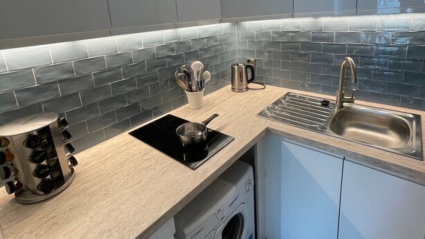 Cocina compacta con armarios grises, una placa de inducción negra, un lavaplatos de acero inoxidable y una lavadora.