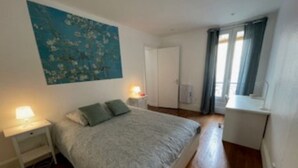 Un dormitorio con cama, mesita de noche, una obra de arte en la pared y una ventana con cortinas.