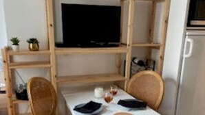 Una habitación con una mesa blanca, dos sillas de mimbre, un televisor sobre una estantería de madera y un refrigerador.