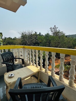 A balcony with a yellow railing, two black chairs, a small table, and a view of greenery and hills in the background.