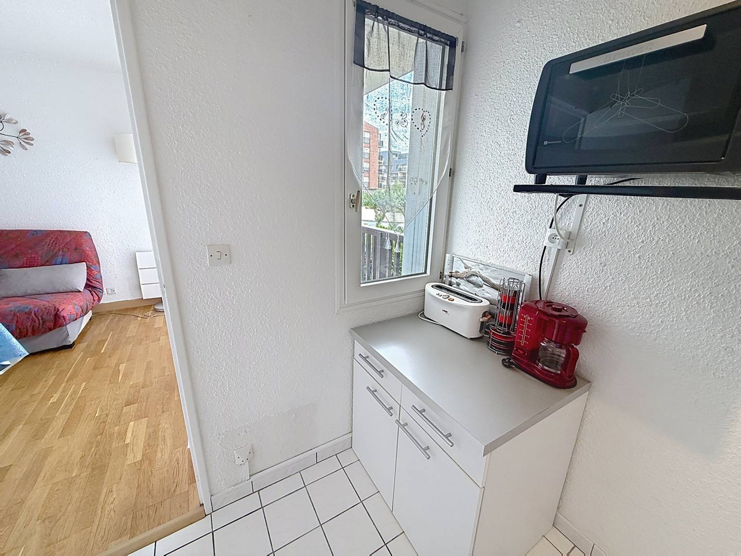 Una cocina compacta con ventana, un tostador, un electrodoméstico rojo y un armario blanco.
