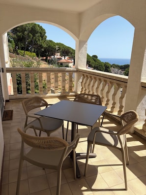 Een overdekt terras met een tafel en stoelen, uitzicht op de zee, en een balkon met een hekwerk.