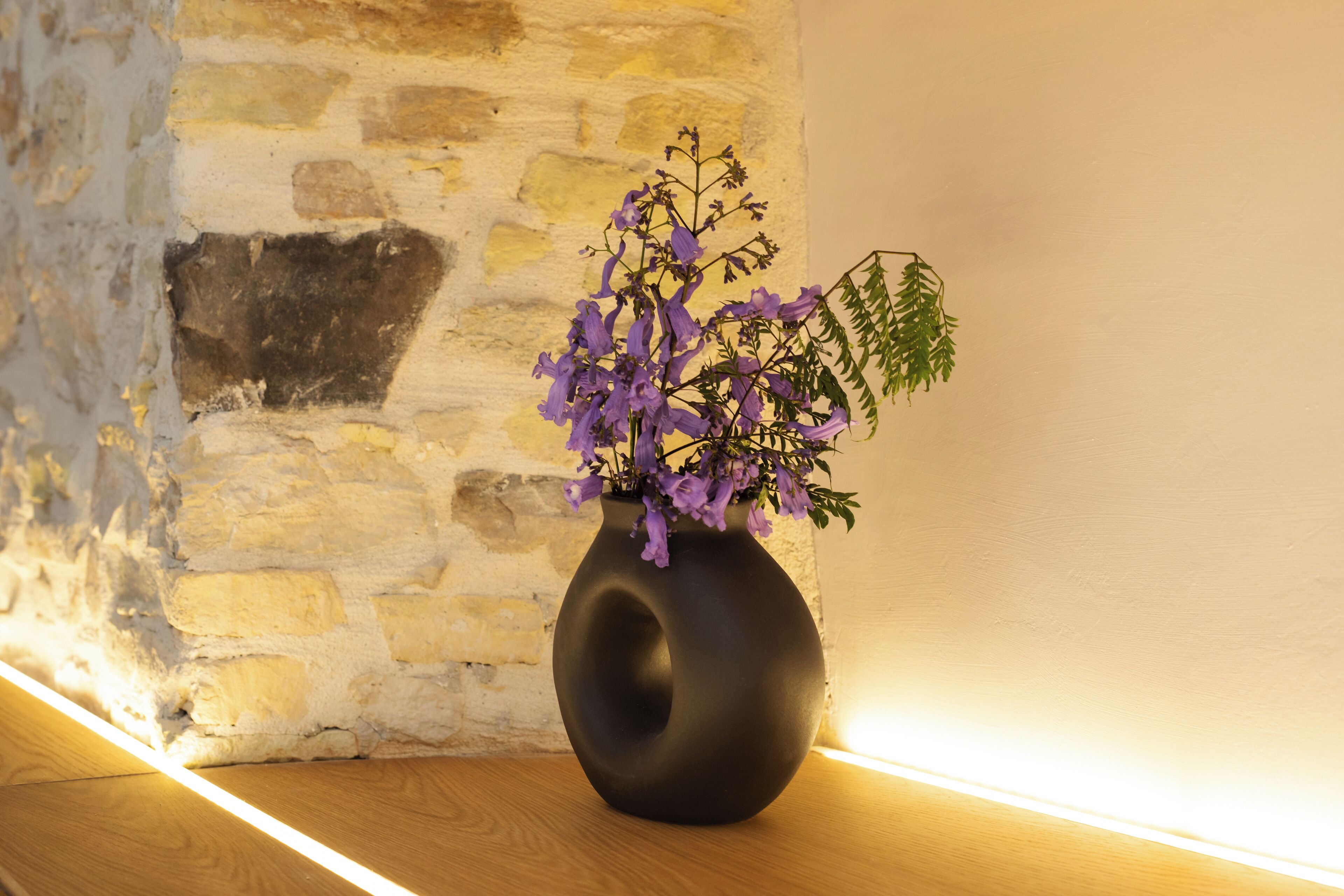 Un vase noir orné de fleurs violettes, posé sur une surface en bois, devant un mur de pierre.